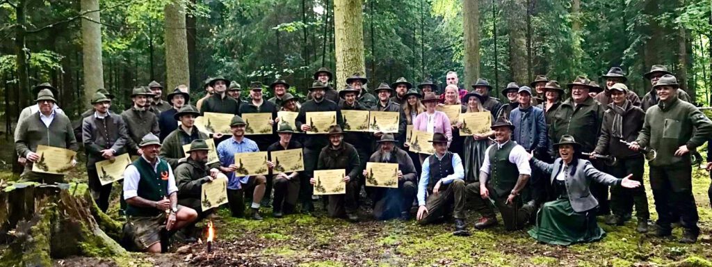 Absolventen der Jagdschule stehen im Wald, schauen in die Kamera und halten ihre Jagdschein-Urkunde hoch